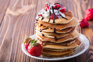  Stack of pancakes with strawberries, whip cream and chocolate syrup on a white plate on a wooden...
