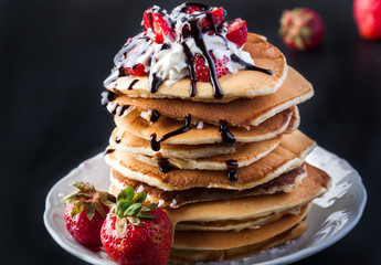  Stack of pancakes with strawberries, whip cream and chocolate syrup on a white plate on a black background. Copy space.