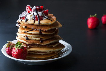 Stack of pancakes with strawberries, whip cream and chocolate syrup on a white plate on a black...