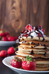 Stack of pancakes with strawberries, whip cream and chocolate syrup on a white plate on a wooden...