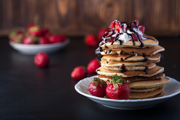 Stack of pancakes with strawberries, whip cream and chocolate syrup on a white plate on a wooden background. Copy space.