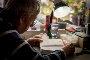 Elderly man with glasses reading writings in notebook near the window at home
