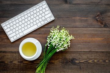 Cosy spring work desk concept. Computer keyboard, cup of tea and bouquet of lily of the valley flowers on dark wooden background top view copy space