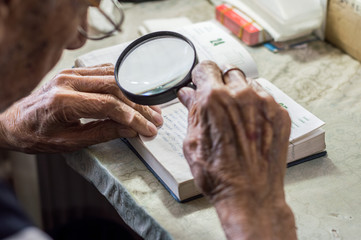 Elderly man reading writings in a notebook with magnifying glass near the window at home