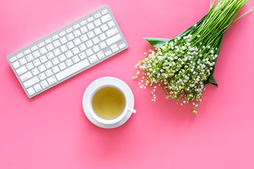 Cosy spring work desk concept. Computer keyboard, cup of tea and bouquet of lily of the valley flowers on pastel pink background top view copy space
