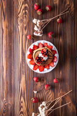 Homemade pancakes with strawberries, whipped cream and chocolate topping, decorated with flowers on a brown wooden background. Overhead shot. Flat lay. Copy space
