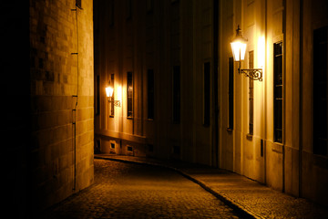 Old lanterns illuminating a dark alleyway medieval street at night in Prague, Czech Republic. Low key photo with brown yellow tones from the lanterns as single light sources against the dark shadows - obrazy, fototapety, plakaty