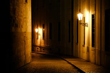 Fotobehang Old lanterns illuminating a dark alleyway medieval street at night in Prague, Czech Republic. Low key photo with brown yellow tones from the lanterns as single light sources against the dark shadows © fewerton