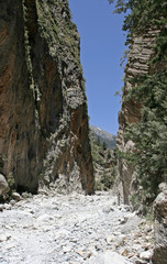 The gorge with high steep rocky slopes with green plants and the small white stony path on its...