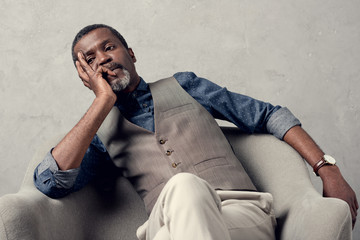 bored stylish african american man in waistcoat sitting in armchair