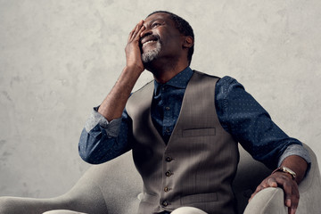 portrait of stylish laughing african american man in waistcoat sitting in armchair