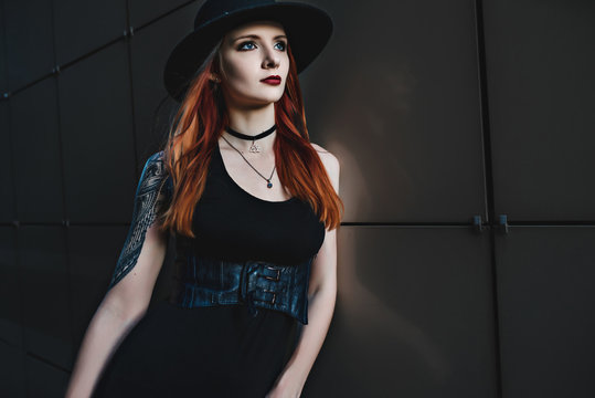 Creative redhead woman in gothic black dress and hat in the city. Street style