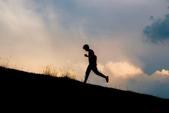 Attractive fit man running fast along big modern bridge at sunset light, black man doing workout outdoors, silhouette runner in windbreaker jogging over bridge road with amazing sunset on background - Powered by Adobe