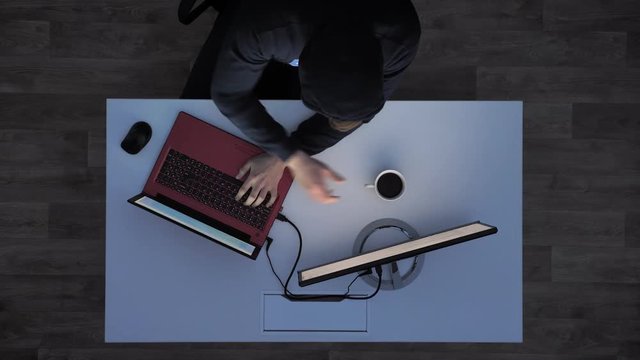 Young man in black hoodie drinking coffee and typing on laptop during night, sitting behind white desk, top shot