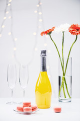 close up view of macarons, empty glasses, bottle of champagne and bouquet of gerbera flowers on grey backdrop