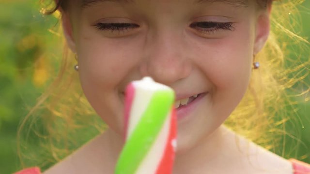 Child in hat licks ice cream. Girl is eating delicious cream ice cream. Ice-cream multicolored on a stick with a strawberry taste is a favorite summer dish of children. Sunset rays shine in the lens.