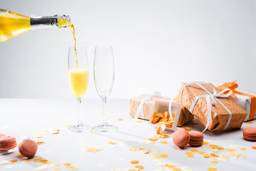 close up view of pouring yellow champagne into glasses process, macarons and arranged gifts on grey backdrop