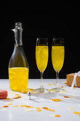 close up view of bottle and glasses of yellow champagne, macaron and gift on surface on black background