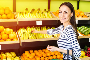 Attractive smiling young girl in casual clothes selects fruits and vegetables from a shelf in a grocery store.