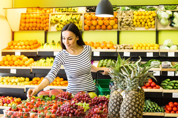 Attractive smiling young girl in casual clothes selects fruits and vegetables from a shelf in a grocery store.