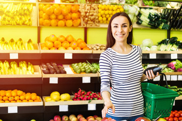Attractive smiling young girl in casual clothes selects fruits and vegetables from a shelf in a grocery store.