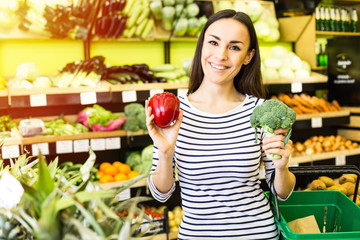 Attractive smiling young girl in casual clothes selects fruits and vegetables from a shelf in a grocery store.
