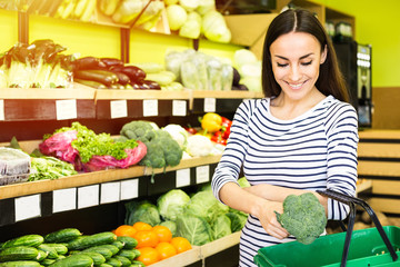 Attractive smiling young girl in casual clothes selects fruits and vegetables from a shelf in a grocery store.
