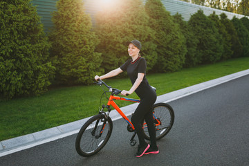 Young athletic brunette strong woman in black uniform, cap stop riding road on black bicycle with orange elements outdoors on spring or summer sunny day. Fitness, sport, healthy lifestyle concept.