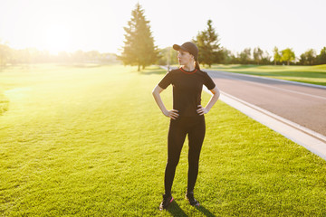 Young athletic girl in black uniform, cap doing sport exercises, warm-up, stretching before running on green lawn in golf course park outdoors on sunny summer day. Fitness, healthy lifestyle concept.