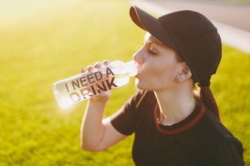 Young girl in black uniform, cap sitting relaxing resting on green lawn after sport exercises, running, drinking water from bottle on outdoors on sunny summer day. Fitness, healthy lifestyle concept.