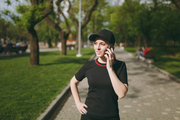 Young athletic beautiful brunette girl in black uniform and cap talking on mobile phone during training, looking aside and standing in city park outdoors. Fitness, healthy lifestyle concept.