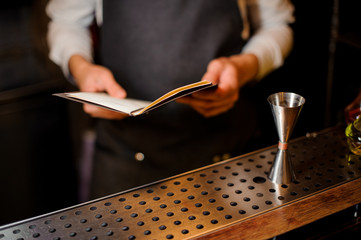 Bartender standing at the bar counter with a recipe book - obrazy, fototapety, plakaty