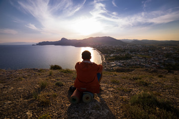 Naklejka premium man sits on top of a hill and looks at the sun set near the sea
