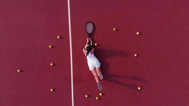 OVERHEAD DRONE shot of young Caucasian teen model wearing fashionable tennis dress, lying on tennis hardcourt with a lot of balls, summer sunny day outdoors. Fashion portrait shoot