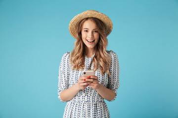 Smiling blonde woman in dress and hat holding smartphone