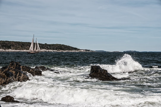 The coast of the Atlantic Ocean. A floating sailboat, stones at the shore amid the waves. The atmosphere of sea voyage, adventurism and romance. USA. Maine.
