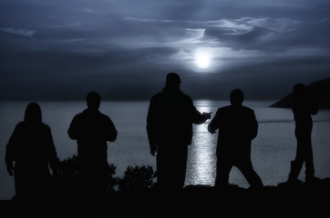 People are staring at the full moon that shines above the sea creating beautiful reflections on the sea