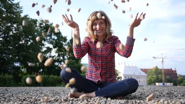Young happy woman enjoy sit on pebbles amuse and smile throw handful of little stones on camera