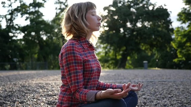 Young woman meditating relaxing sitting in a park on glade pebbels ground in sunset sun rays