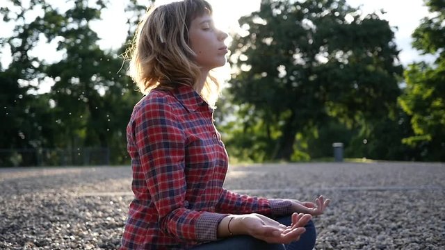 Young woman meditating relaxing sitting in a park on glade pebbels ground in sunset sun rays