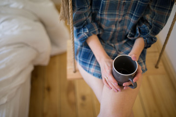 Beautiful girl is sitting on swing in loft minimalist interior. Young woman in men's checkered shirt is drinking coffee. Cozy morning photo.