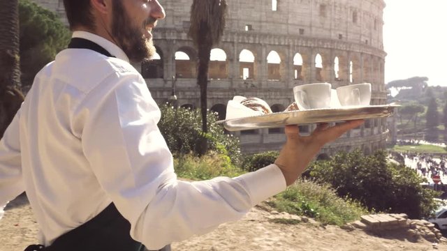 Elegant smiling waiter serving breakfast with breakfast coffee croissant cappuccino on a try outdoor to a bar restaurant table in front of colosseum in rome at sunset