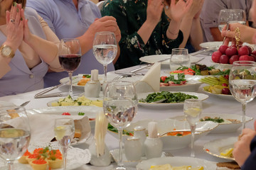 People put their hands together or clap their hands. Close-up of a festive table. Red wine and vodka are poured into the glasses. On the table are dishes with fruits, salads and snacks. Life style.