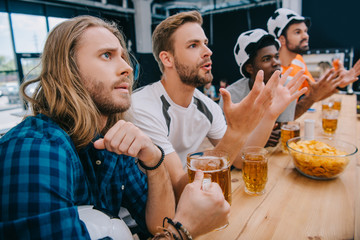 upset multicultural group of male football fans in soccer ball hats drinking beer and watching football match at bar