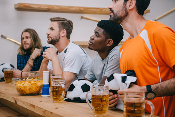 multicultural group of male football fans with soccer ball hats, fan horn, chips and beer watching soccer match at bar