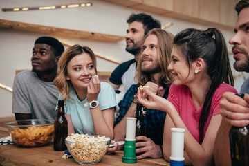 multiethnic group of friends watching football match at bar with fan horns, chips, popcorn, beer and soccer ball