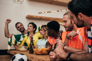 group of multicultural friends in fan t-shirts with beer bottles watching soccer match at bar