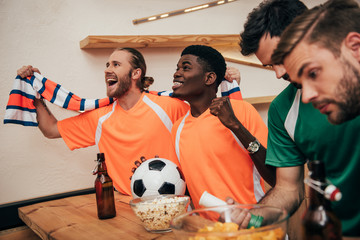 smiling multicultural football fans in orange t-shirts and scarf celebrating victory with ball while their upset friends in different t-shirts sitting near during watch of soccer match at bar