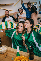 high angle view of women in green fan t-shirts holding fan scarf and their male friends standing behind during watch of soccer match at bar