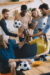 group of multicultural friends with soccer ball clinking by beer bottles during football match at...